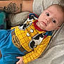 baby, child, person, tattoo, arm, clothing, cowboy_outfit, yellow_shirt, blue_pants, indoor, sofa, couch, casual, portrait, infant, cute, relaxing, human, face, expression