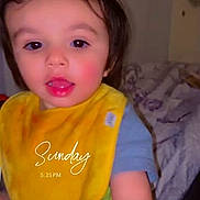 Braelynn joined the competition — help win amazing prizes! toddler, child, yellow_bib, indoor, bed, blanket, face, person, sitting, curious, short_hair, blue_shirt, skin, mouth_open, eyes, photography, portrait, music_overlay, timestamp, casual
