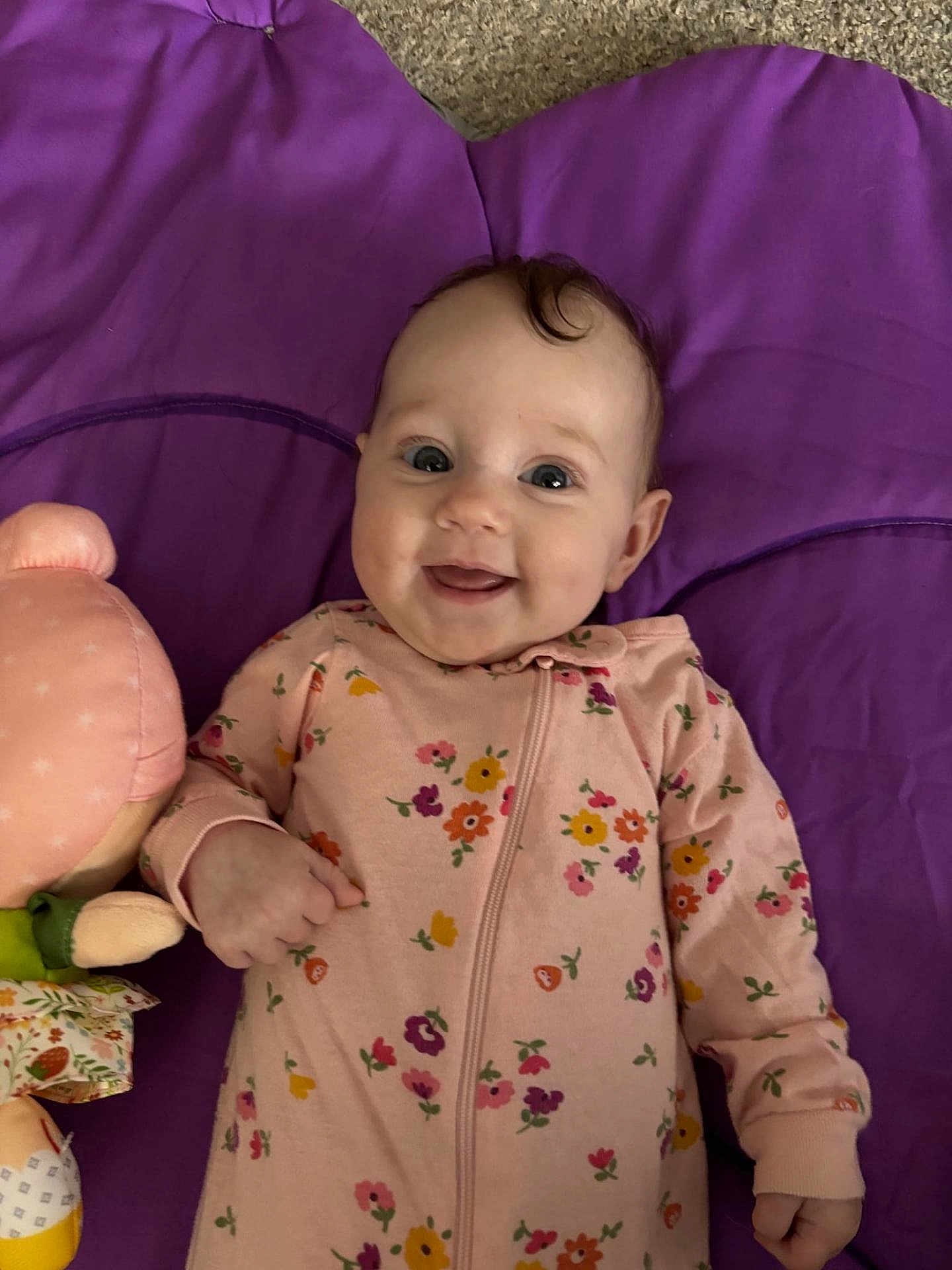 Everly is registered to the contest to win money with this photo: baby, infant, smiling, face, eyes, pajamas, floral_pattern, purple_mat, plush_toy, hand, cheek, portrait, laying_down, head, skin, cute, happy, carpet, soft_toy, nursery