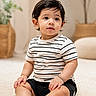 toddler, child, baby, striped_tshirt, black_shorts, sitting, carpet, indoor, home_decor, plant, curly_hair, cute, portrait, soft_light, natural_light, expression, furniture, floor, skin, smile