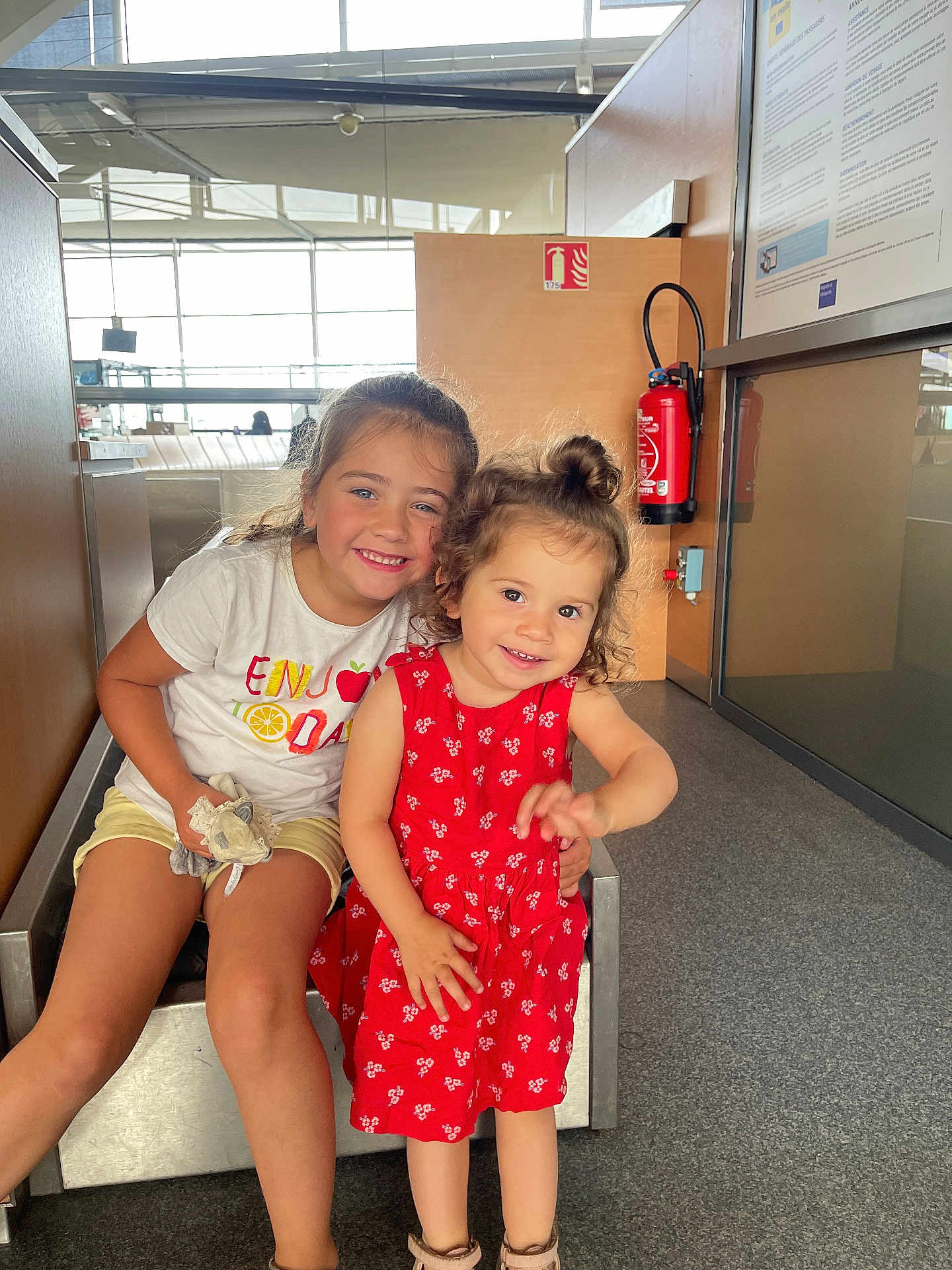 Margot participe au concours pour gagner de l'argent avec cette photo : child, children, girl, smile, happy, indoor, red_dress, white_shirt, toy, curly_hair, long_hair, modern_interior, floor, bench, window, fire_extinguisher, casual_clothing, playful, cute, portrait