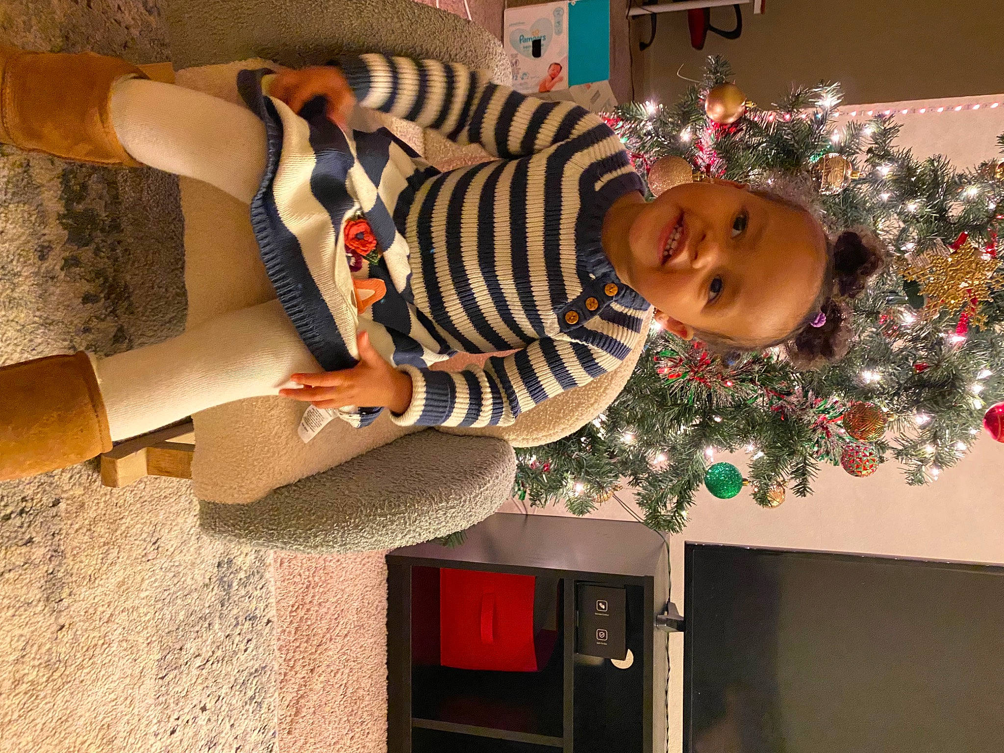 Leia is registered to the contest to win money with this photo: christmas_decoration, christmas_ornament, christmas_tree, event, happy, holiday, holiday_ornament, human_leg, joy, knee, person, plant, red, sleeve, smile, standing, thigh, toddler, tree, waist