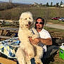 man, dog, fluffy_dog, picnic_table, quilt, outdoor, vineyard, grass, trees, sunlight, sunglasses, cooler, fire_pit, cars, rural, nature, relaxation, casual, daytime, scenery