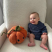 Colton is registered to the contest to win money with this photo: baby, child, plush_pumpkin, armchair, cozy, smiling, indoor, onesie, cute, soft_texture, orange, cream_color, happy, sitting, toy, infant, cushion, decor, comfort, face