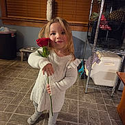 Athena is registered to the contest to win money with this photo: child, girl, rose, flower, white_sweater, long_hair, indoor, floor_tiles, window_blinds, smile, person, standing, warm_lighting, kitchen_shelf, pineapple, cozy, cute, holding_flower, home, casual_clothing