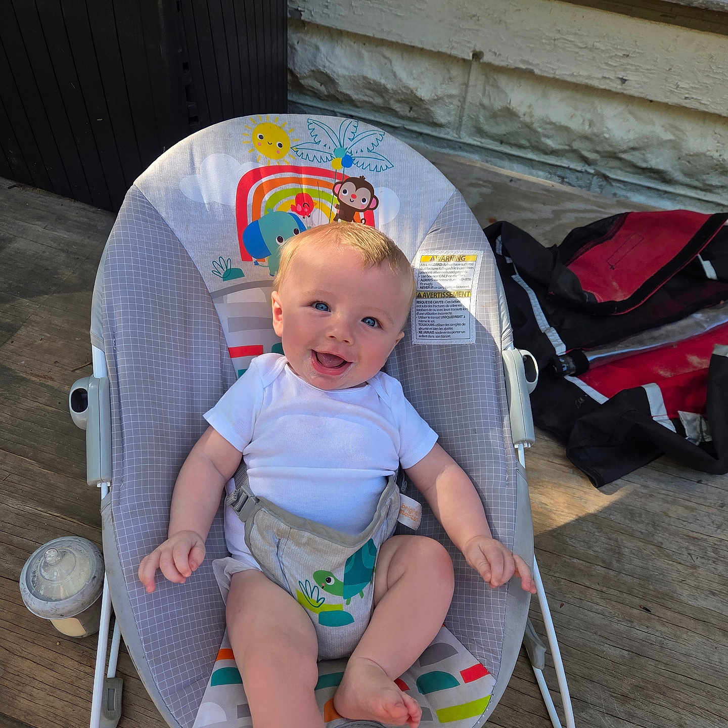 Caspian is registered to the contest to win money with this photo: baby, infant, smile, baby_rocker, blue_eyes, white_onesie, wooden_deck, baby_bottle, outdoor, sunlight, happy, child, seat, toy_pattern, casual, playful, colorful, comfort, person, daylight
