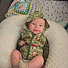 Caspian is registered to the contest to win money with this photo: baby, infant, camo_hoodie, cushion, blanket, playpen, toy, smile, socks, cute, child, indoor, soft_texture, happy, comfort, clothing, face, person, cozy, resting