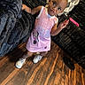 toddler, child, pink_dress, minnie_mouse, white_sneakers, wooden_floor, black_couch, pillow, indoor, standing, curious_expression, hair_bow, floor, furniture, person, clothing, young_child, cute, home, casual