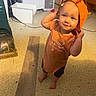 toddler, child, orange_clothing, barefoot, indoor, floor, vacuum_cleaner, hat, smile, curious, standing, person, young_child, casual_wear, headwear, home, messy, toy, expression, flooring