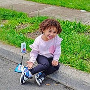 Miracle is registered to the contest to win money with this photo: casual_clothing, child, curb, curly_hair, daylight, fence, grass, happy, house, juice_box, nature, outdoor, playground, residential_area, sidewalk, sitting, smiling, snack, sneakers, trees