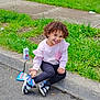 child, curly_hair, smiling, sitting, curb, street, grass, juice_box, snack_bag, sneakers, pink_sweater, black_leggings, outdoor, daytime, residential_area, deflated_inflatable, happy, casual, person, sidewalk