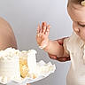 baby, child, cake, frosting, hand, adult, support, white, plate, celebration, food, dessert, curious, messy, closeup, portrait, indoor, light, holding, cute