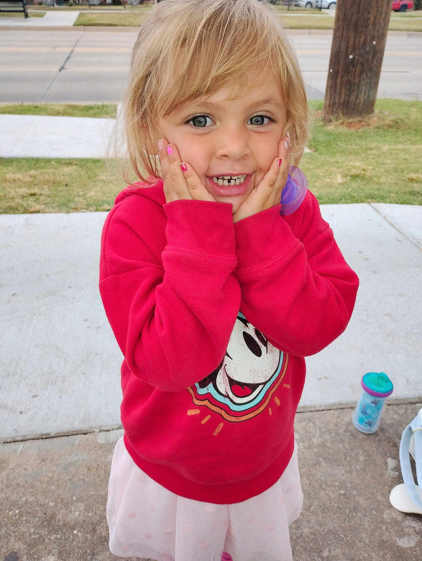 Brandy is registered to the contest to win money with this photo: child, girl, red_sweatshirt, cartoon_character, smiling, hands_on_face, outdoor, sidewalk, grass, road, pacifier, sippy_cup, blonde_hair, blue_eyes, happy, casual_clothing, playful, portrait, cute, young