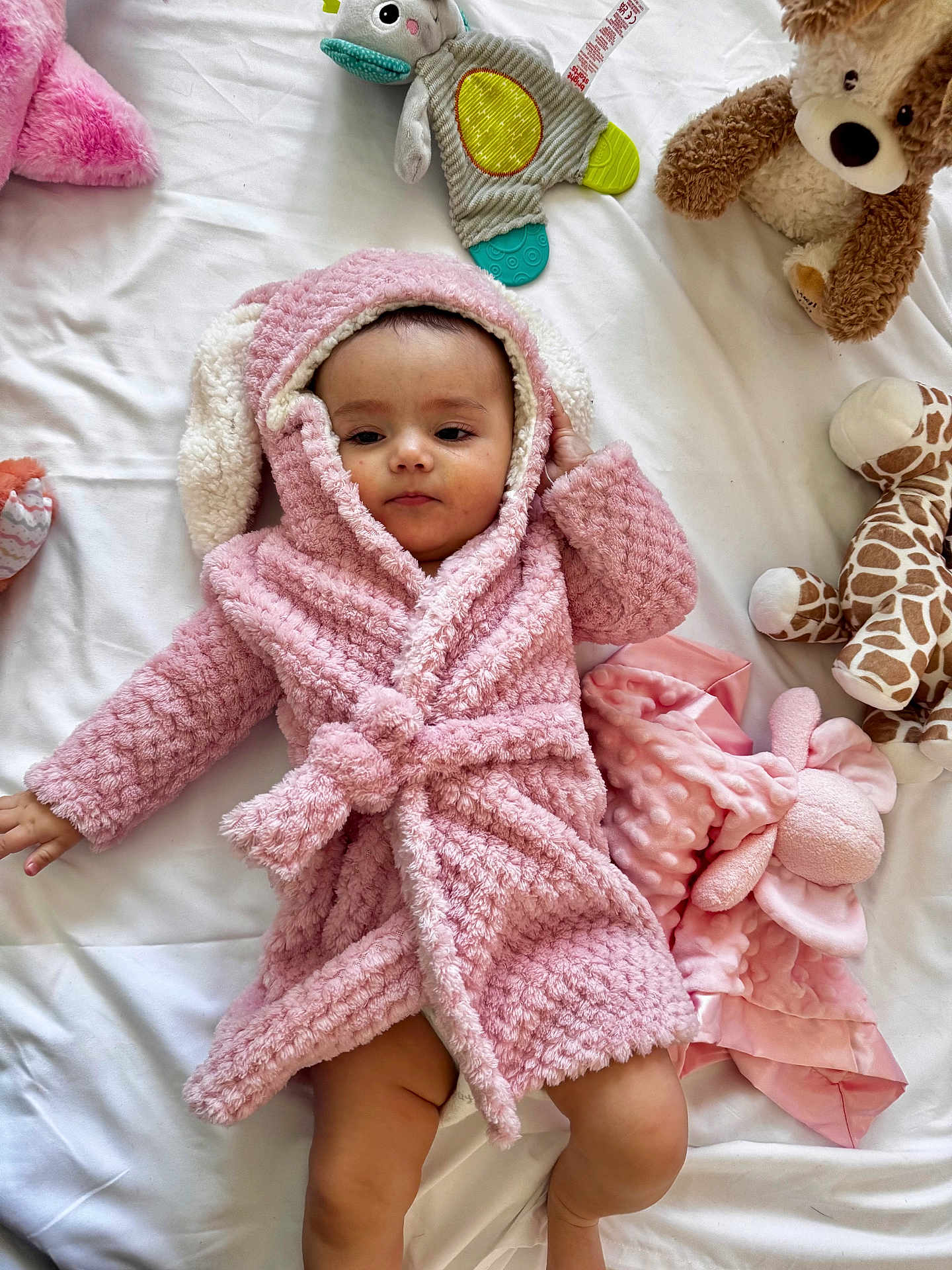 Mayah is registered to the contest to win money with this photo: baby, pink_robe, bunny_ears, plush_toys, giraffe_toy, blanket, bed, soft_toys, child, infant, cute, indoors, sleepwear, toddler, comfort, cuddly, white_bed, relaxed, cozy, warm