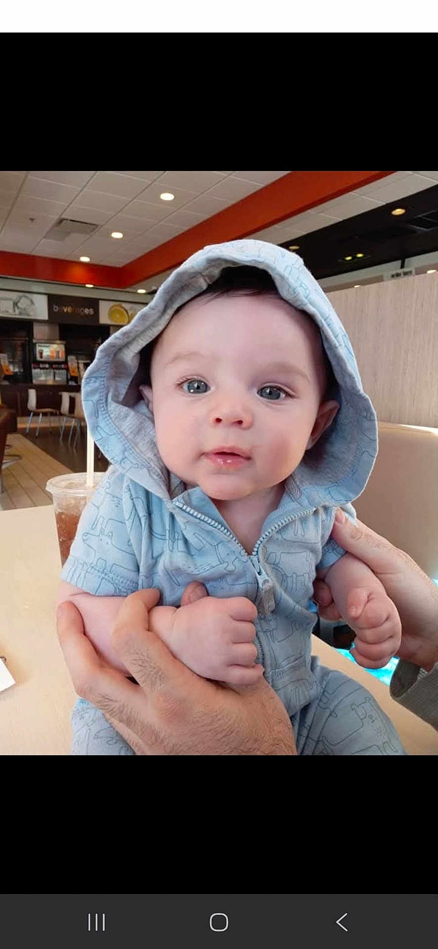 Daniel joined the competition — help win amazing prizes! baby, child, hoodie, blue_clothing, hands, face, eyes, indoor, restaurant, table, drink, cup, person, seated, casual, portrait, cute, infant, holding, expression
