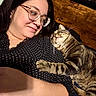 affection, casual, cat, close_up, face, glasses, human, indoor, intimate, lying_down, paw, pet, polka_dot, relaxed, resting, striped_fur, tabby_cat, warm, woman, wooden_background