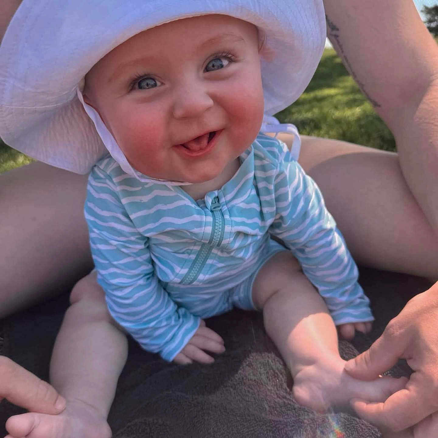 Grayson is registered to the contest to win money with this photo: baby, blue_clothing, child, cute, daylight, grass, hands, happy, hat, infant, legs, outdoor, playful, portrait, sitting, skin, smiling, striped_clothing, sunhat, towel