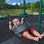 Easton is registered to the contest to win money with this photo: baby, child, swing, playground, outdoor, red_shorts, grass, trees, sky, clouds, metal_chains, black_rubber_surface, green_poles, barefoot, infant, summer, fun, park, seat, daytime
