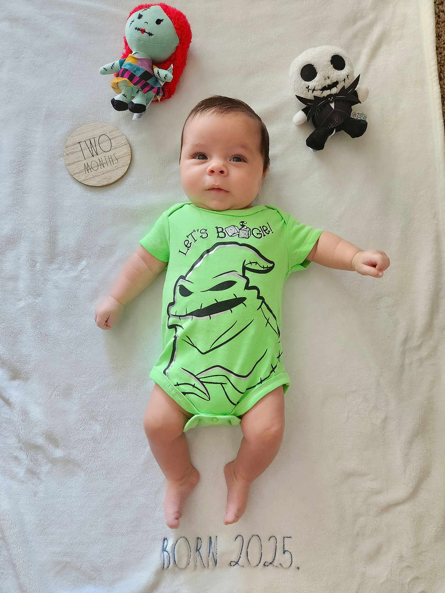 Austin is registered to the contest to win money with this photo: baby, infant, onesie, green_clothing, plush_doll, skull_doll, red_hair_doll, blanket, milestone, wooden_plaque, text, cute, lying_down, infant_face, portrait, celebration, soft_texture, child, two_months, embroidered_text