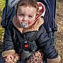 child, baby, toddler, pacifier, stroller, jacket, fur_lined_hood, harness, buckle, patterned_leggings, leopard_print, seated, outdoors, park_path, smiling, blue_eyes, hand, brown_hair, sunlight, gravel_path