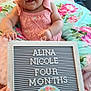 baby, infant, smile, pink_clothing, headband, bow, letter_board, hand, pillow, floral_pattern, blanket, celebration, milestone, cute, child, portrait, sitting, happy, indoors, person
