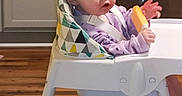Maria is registered to the contest to win money with this photo: baby, high_chair, popsicle, child, indoor, kitchen, flooring, cabinet, appliance, seat, person, clothing, curious, orange_food, young_child, face, hair, wood_floor, table, cushion