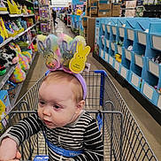 Maria is registered to the contest to win money with this photo: baby, child, shopping_cart, striped_clothing, bunny_ears, store_aisle, toys, holiday_decorations, hand_holding, retail_store, colorful, infant, headwear, curious, indoor, person, shopping, seat_belt, floor, shelves