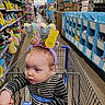baby, child, shopping_cart, striped_clothing, bunny_ears, store_aisle, toys, holiday_decorations, hand_holding, retail_store, colorful, infant, headwear, curious, indoor, person, shopping, seat_belt, floor, shelves
