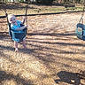 Maria is registered to the contest to win money with this photo: baby, swing, playground, child, outdoor, sunlight, woodchips, bench, trees, smiling, happy, blue_clothing, person, daylight, nature, shadow, grass, seat, park, play