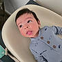 baby, child, infant, onesie, face, tongue_out, curious, blue_clothing, airplane_print, cloud_print, baby_chair, indoor, carpet, table, strap, wood_furniture, cute, lying_down, portrait, soft_lighting