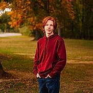Elijah is registered to the contest to win money with this photo: red_hair, teenager, hoodie, maroon_hoodie, jeans, hands_in_pockets, portrait, outdoor, park, autumn, fall_leaves, tree, grass, freckles, standing, young_person, casual_clothing, smiling, bokeh, natural_light