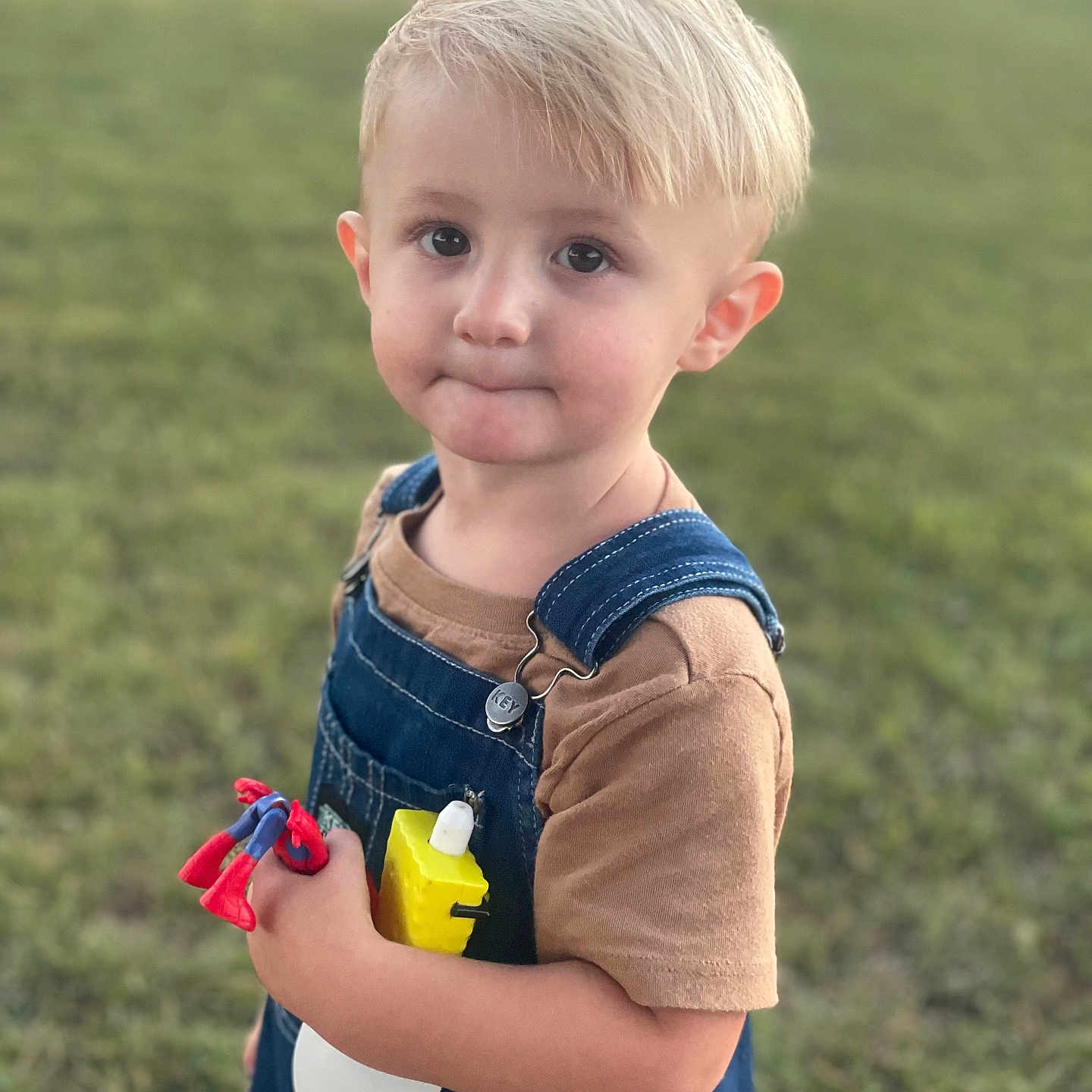 Oscar Felix is registered to the contest to win money with this photo: blonde_hair, boy, brown_tshirt, casual_clothing, child, cute, daylight, denim_overalls, grass, innocent, nature, outdoor, person, playtime, portrait, spiderman, spray_bottle, toddler, toy, young_child