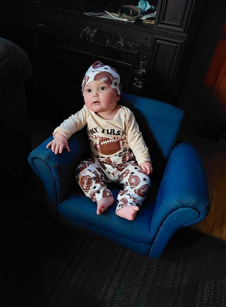 Austin is registered to the contest to win money with this photo: baby, child, infant, blue_chair, football_pajamas, hat, indoor, portrait, curious, seated, wooden_floor, cozy, cute, wide_eyes, small_chair, pajamas, clothing, person, furniture, young_child