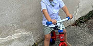 Manel a rejoint le concours — aidez-le/la à gagner de superbes lots ! balance_bike, boy, casual_clothing, child, fun, happy, outdoor, pavement, play, portrait, sandals, shirt, shorts, smiling, standing, summer, toy, vehicle, wall, young
