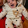 baby, child, smiling, happy, onesie, polka_dots, bear_patch, hooded_clothing, indoor, person_holding, red_clothing, face, expression, warm_clothing, floor, wooden_floor, infant, cute, portrait, cozy
