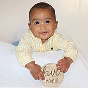 Caden is registered to the contest to win money with this photo: baby, infant, child, smiling, face, hands, shirt, striped_clothing, yellow, wooden_sign, text, five_months, portrait, cute, indoors, white_background, happy, young_baby, person, sitting