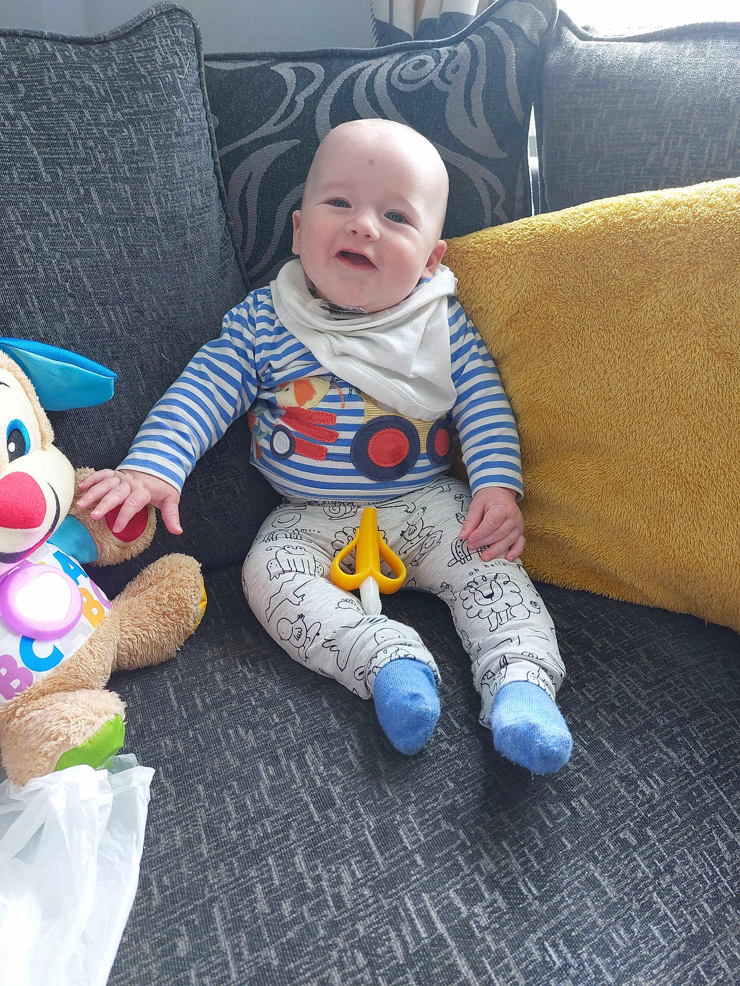 Carter-Jack is registered to the contest to win money with this photo: baby, child, smiling, couch, cushion, toy, plush_toy, striped_clothing, patterned_pants, blue_socks, indoor, happy, seated, person, fabric, living_room, yellow_pillow, white_bib, playful, scissors_toy