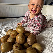 Eevy participe au concours pour gagner de l'argent avec cette photo : toddler, child, kiwi, fruit, bed, white_bedspread, smiling, happy, indoor, floral_clothing, pile, food, texture, natural_light, person, cute, playful, skin, head, expression