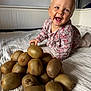 toddler, child, kiwi, fruit, bed, white_bedspread, smiling, happy, indoor, floral_clothing, pile, food, texture, natural_light, person, cute, playful, skin, head, expression