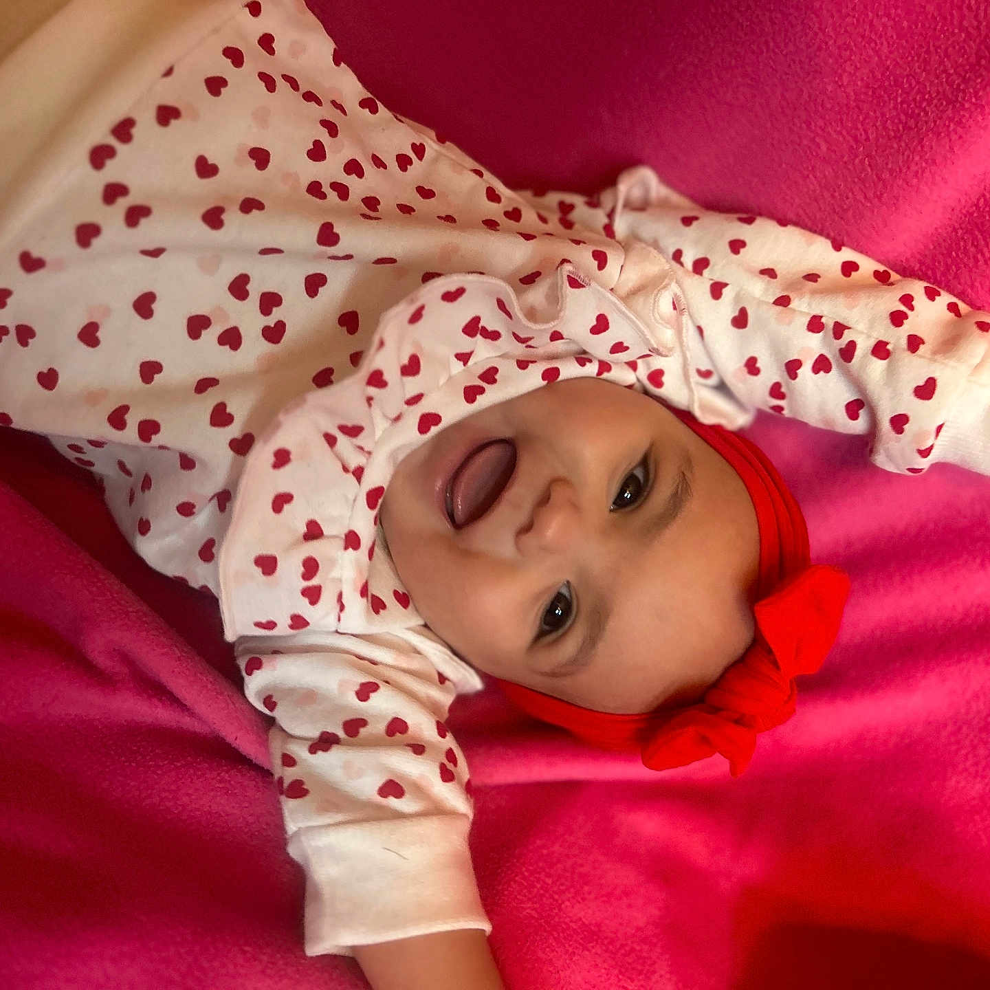 Kaileeanna is registered to the contest to win money with this photo: baby, infant, smiling, red_headband, heart_pattern, white_clothing, pink_blanket, happy, cute, child, portrait, lying_down, indoors, playful, face, person, toddler, headwear, expression, joy
