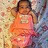 baby, infant, child, floral_blanket, coral_outfit, smiling, cute, indoors, lying_down, hair_clip, text_on_clothing, skin, legs, arm, hand, fabric, pattern, comfortable, happy, portrait