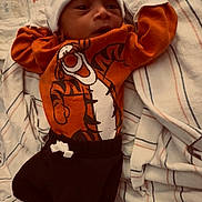 Zyel joined the competition — help win amazing prizes! baby, black_pants, blanket, casual_clothing, child, cozy, cute, face, headwear, indoors, infant, lying_down, newborn, orange_clothing, person, portrait, soft_lighting, striped_blanket, tiger_shirt, white_hat