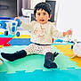 toddler, child, toy, playmat, indoor, smiling, sitting, colorful, car_toy, leggings, socks, long_sleeve_shirt, black_hair, face, young_child, floor, playtime, cute, happy, home