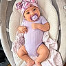 baby, infant, child, portrait, lying_down, pacifier, hat, bow, floral, lavender, purple, onesie, blanket, cushion, baby_seat, indoor, cozy, smiling, face, feet