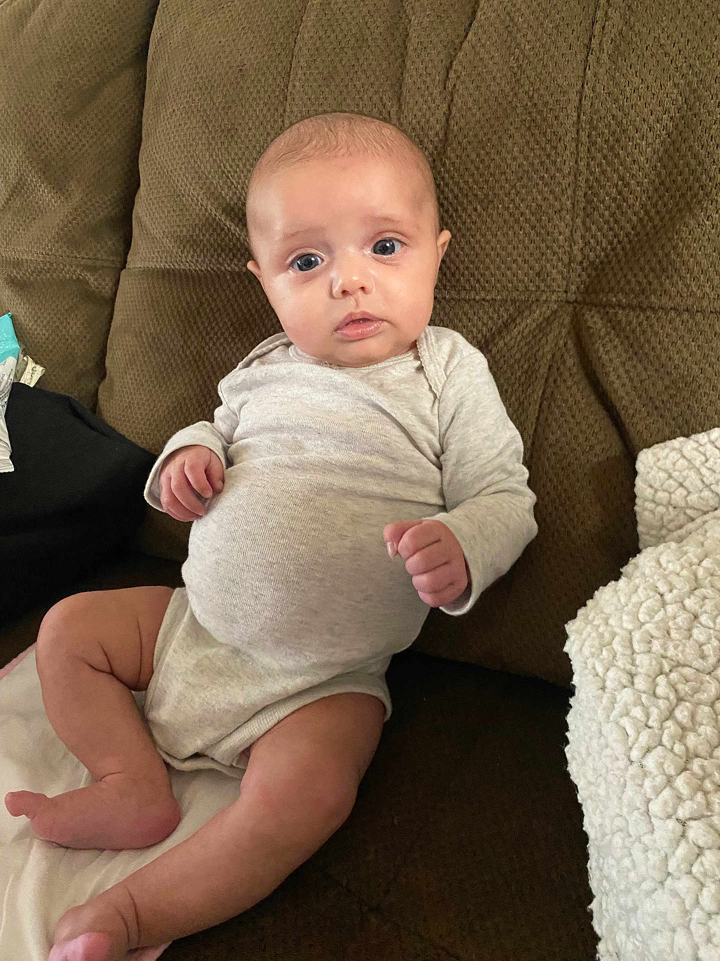 Mckenna is registered to the contest to win money with this photo: baby, infant, couch, blanket, onesie, indoor, person, face, blue_eyes, skin, hand, foot, leg, fabric, texture, soft, seated, looking, curious, cozy