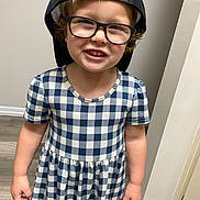 Sailor is registered to the contest to win money with this photo: baseboard, black_cap, cap, checkered_dress, child, curly_hair, dress, floor, glasses, happy, indoor, light_skin, person, plaid, short_sleeves, smile, standing, teeth, wall, young_child