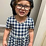 baseboard, black_cap, cap, checkered_dress, child, curly_hair, dress, floor, glasses, happy, indoor, light_skin, person, plaid, short_sleeves, smile, standing, teeth, wall, young_child