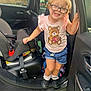 child, girl, glasses, pink_bow, teddy_bear_shirt, denim_shorts, frilly_socks, black_shoes, car_interior, car_door, car_seat, child_seat, smiling, standing, holding, outdoor, grass, fence, daylight, cute