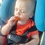 baby, child, car_seat, orange_shirt, snack, messy_face, black_harness, seatbelt, closed_eyes, eating, hand, car, window, seat, infant, young_child, cute, food, mess, portrait