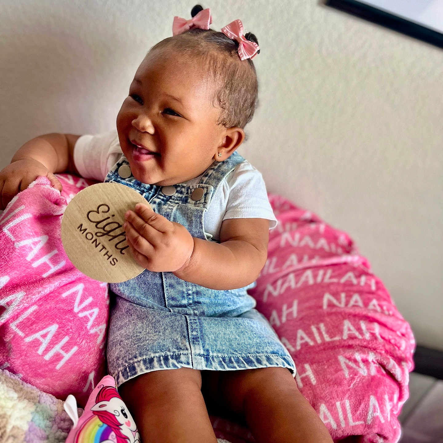 Naailah joined the competition — help win amazing prizes! baby, bracelet, child, cozy, cushion, cute, denim_dress, eight_months, girl, happy, indoors, personalized_blanket, pink_bows, portrait, sitting, smiling, toy, unicorn, white_shirt, wooden_sign